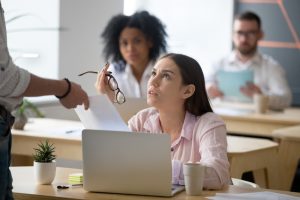 Mulher sentada à mesa de trabalho olha de modo desconfortável para um colaborador em pé que aponta o dedo em riste