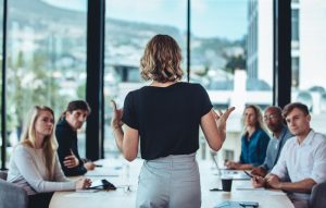 mulher de costas para a câmera e conversando com quatro pessoas numa mesa de reunião