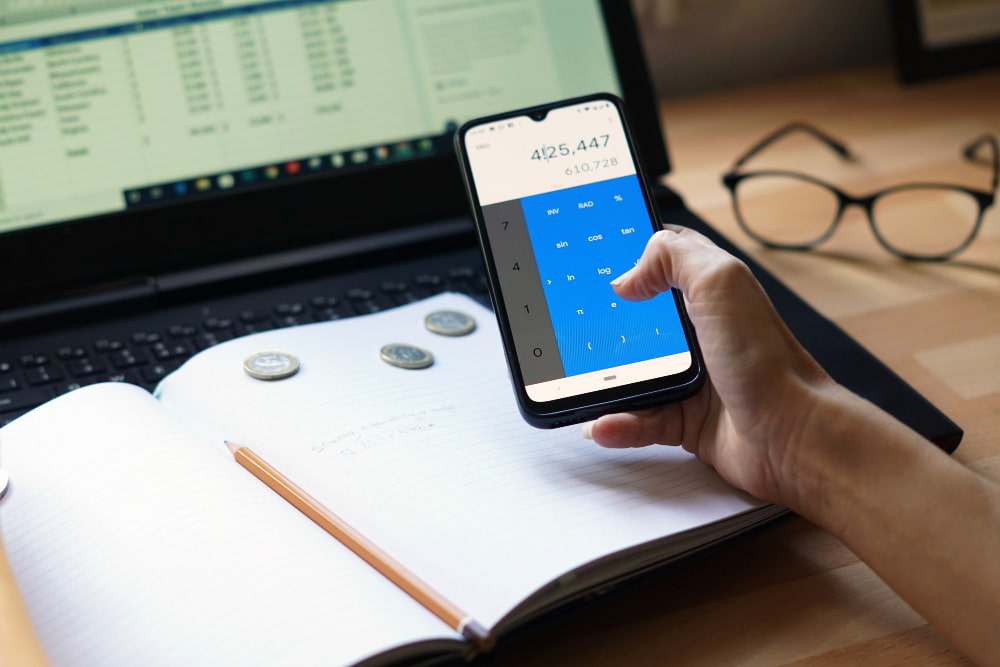 pessoa segura um celular com a calculadora aberta sobre uma mesa com notebook e um caderno com lápis e moedas em cima.