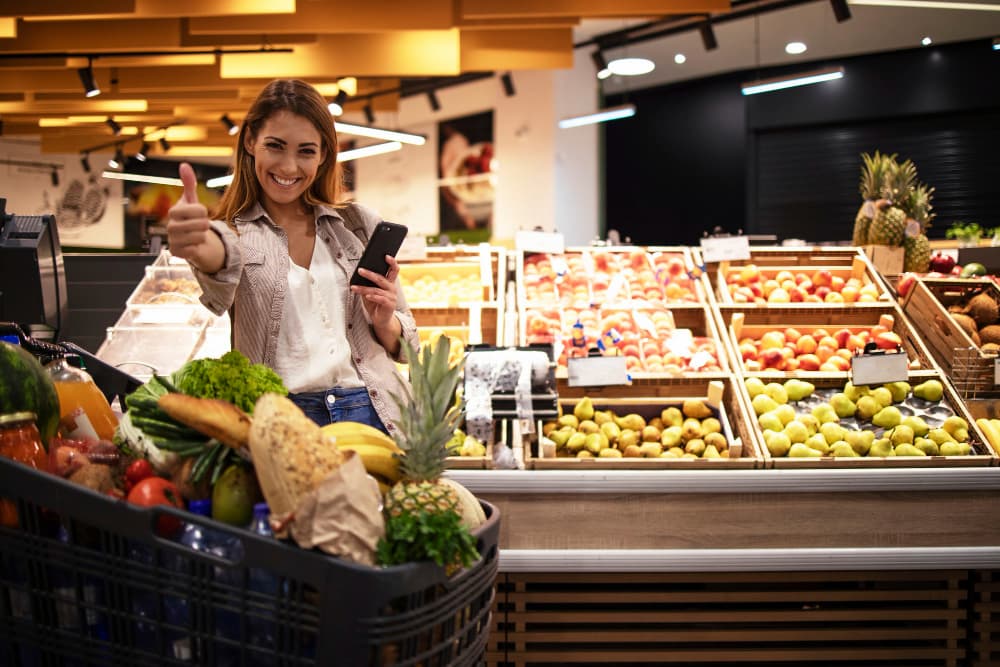 mulher no supermercado em frente à banca de frutas e seu carrinho de compras segura seu smartphone enquanto faz sinal positivo com as mãos.