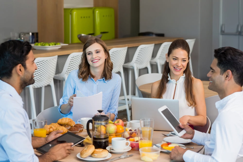 grupo de quatro pessoas composto por dois homens e duas mulheres trabalham reunidos em uma mesa redonda enquanto tomam café da manhã
