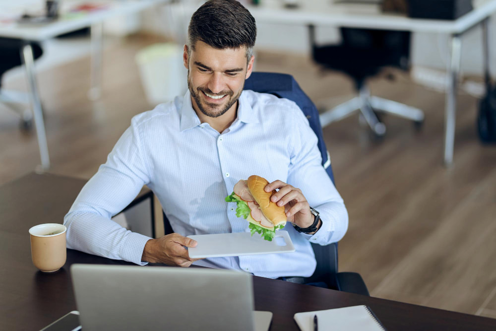 colaborador sorridente comendo sanduíche enquanto utiliza um computador no escritório