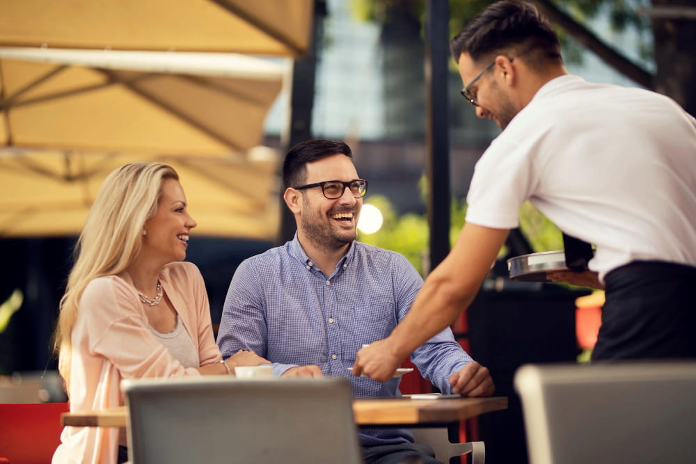 homem e mulher sorridentes sentados em uma mesa de restaurante são servidos por um garçom