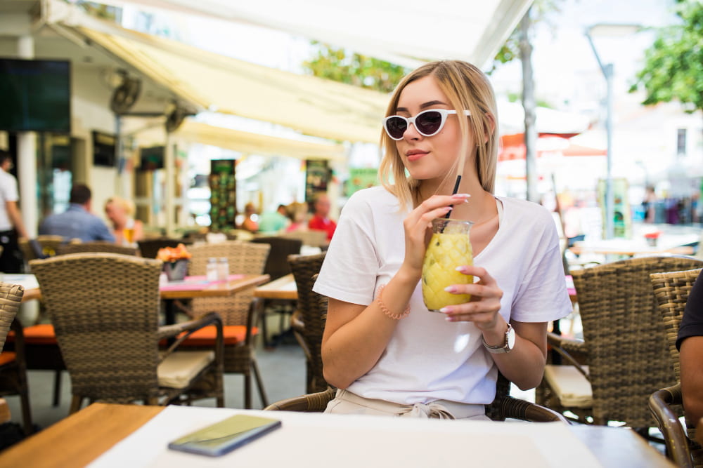 mulher com óculos de sol sentada em uma mesa de restaurante segura um copo de bebida