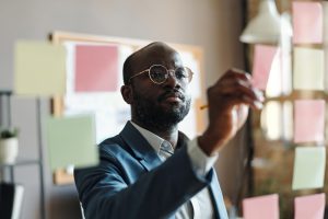 homem em pé escrevendo num post-it pregado em mural de vidro 