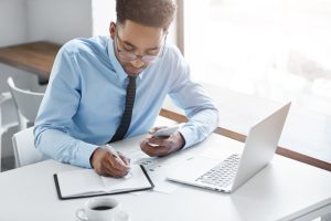 Homem sentado faz anotações em seu caderno em frente ao seu lap top