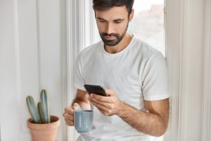 Homem em pé usando celular com caneca na mão direita