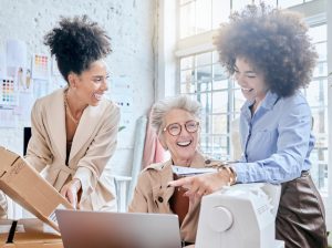 Uma mulher idosa sentada à frente de seu laptop e duas mulheres negras ao seu lado conversando animadamente 