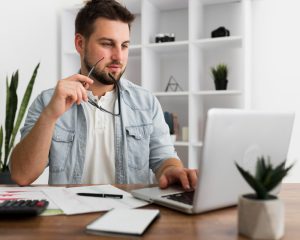Homem sentado à mesa do home office em frente ao seu laptop segurando um óculos e planejando sua carta de apresentação