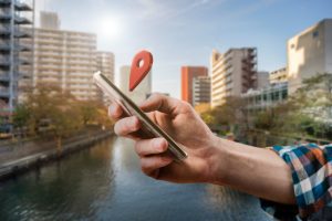 A mão de um homem segura um celular com um pin em 3d de geolocalização com uma cidade e um rio ao fundo