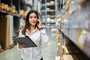 Mulher anda pelo estoque de uma empresa com uma prancheta na mão fazendo auditoria dos produtos e falando ao celular