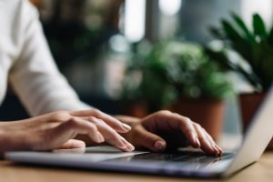 braços de uma pessoa digitando em laptop sobre uma mesa de madeira com plantas desfocadas no fundo