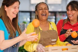 duas mulheres sorridentes brincam com laranjas que outra mulher carrega em cesta