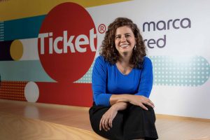 Imagem de Tatiana Romero, diretora de recursos humanos da Ticket. Ela está sentada em frente a um painel da Ticket