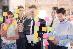 Cunco pessoas em frente a um painel de vidro com post-its colados analisam como implementar a matriz de competências 