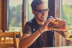 Homem sorridente come um harmburguer, simbolizando hábitos de alimentação nocivos à saúde