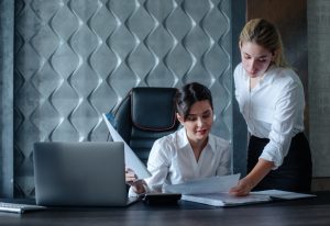 Mulher sentada à mesa de trabalho com seu computador ao lado conversa com mulher em pé que aponta para um papel sobre a mesa