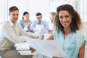 Mulher com papel na mão em primeiro plano e equipe de RH ao fundo fazem planejamento tático