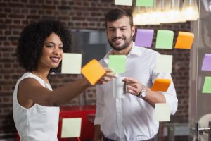 Uma mulher e um homem em pé estão conversando à frente de um painel com post-its colados