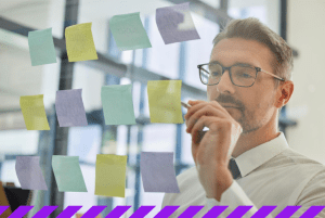 Homem em pé escreve em post-it fixado em painel de vidro o seu planejamento estratégico