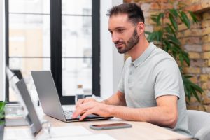 Homem sentado à mesa de trabalho digita em seu laptop