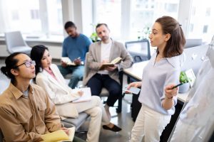 4 pessoas sentadas em ambiente profissional escutam preleção de mulher em pé