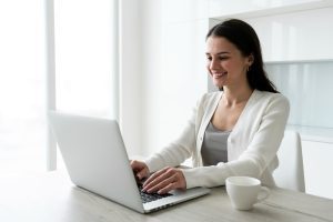 Mulher sentada à mesa de trabalho digita sorridente em laptop