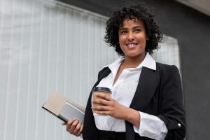 Mulher em pé sorrindo e segurando caderno e café 