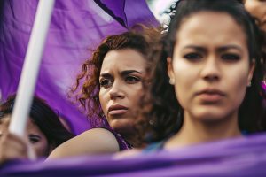 Mulheres empunham bandeiras lilases com expressões graves, simbolizando a luta pelos direitos das mulheres no mercado de trabalho