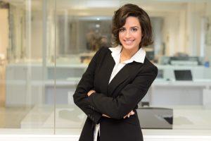 Mulher com roupa executiva em pé de braços cruzados em frente a painel de vidro com escritório ao fundo mostra como melhorar a comunicação no ambiente de trabalho por meio da comunicação não verbal