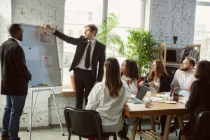 Sala de reuniões com pessoas sentadas e duas em pé mostrando informações em flipchart