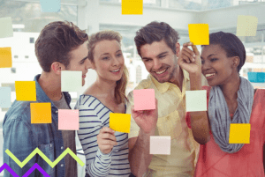 Equipe multifuncional composta por 4 pessoas olham para painel transparente com post-its colados