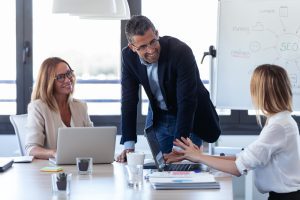 Um homem em pé debruça-se em mesa entre duas mulheres sentadas à mesa de trabalho para conversar amigavelmente mostrando como melhorar a comunicação no ambiente de trabalho facilita as relações