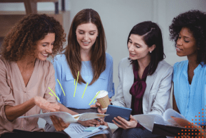 Quatro mulheres em pé observando papeis e sorrindo representando as mulheres no mercado de trabalho
