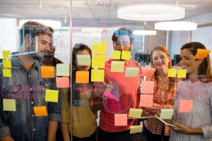 Cinco pessoas em pé olham para painel transparente com post-its colados à sua frente enquanto conversam