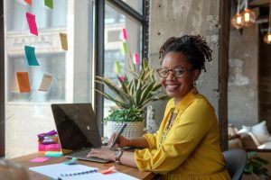 Mulher sorrindo sentada a mesa do trabalho segurando caneta e com mão no teclado