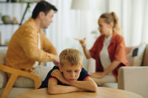 criança triste apoiada na mesa com pais discutindo ao fundo