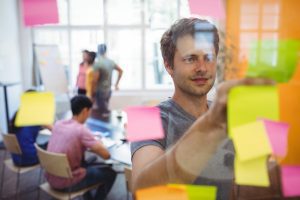 homem cola post-it no painel de vidro enquanto integrantes da equipe trabalham de modo colaborativo em design thinking