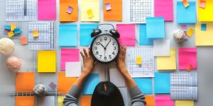 Mulher segura relógio analógico sobre uma mesa com diversos post-its, planners e calendários simbolizando a organização das rotinas contábeis