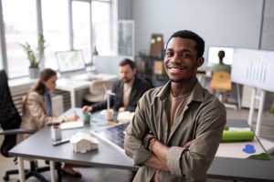 Funcionário sorridente olha para a câmera enquanto colegas trabalham ao fundo demonstrando engajamento por ser ouvido em pesquisa de satisfação
