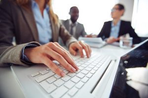 Foto em primeiro plano do teclado de um laptop com as mãos de uma profissional digitando em segundo plano