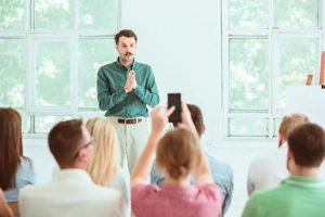 homem em pé com as mãos juntas fazendo workshop com equipe sentada de frente para ele