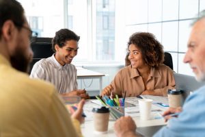 Quatro pessoas, duas em primeiro plano, mais preocupadas e duas em segundo plano, sorridentes, trabalham juntas com resiliência profissional em mesa de escritório