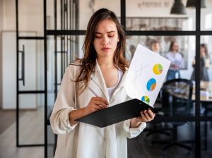 Mulher jovem, vestindo uma camisa clara, segurando um prancheta e analisando gráficos com diferentes tipos de diagramas criando estratégias de employer branding