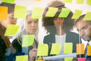 Grupo de quatro pessoas observa e discute anotações em post-its colados em um vidro vivenciando segurança psicológica no trabalho