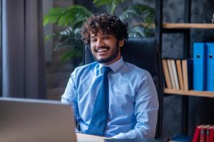 Jovem talento com roupa corporativa sorri para a câmera sentado em sua mesa de trabalho