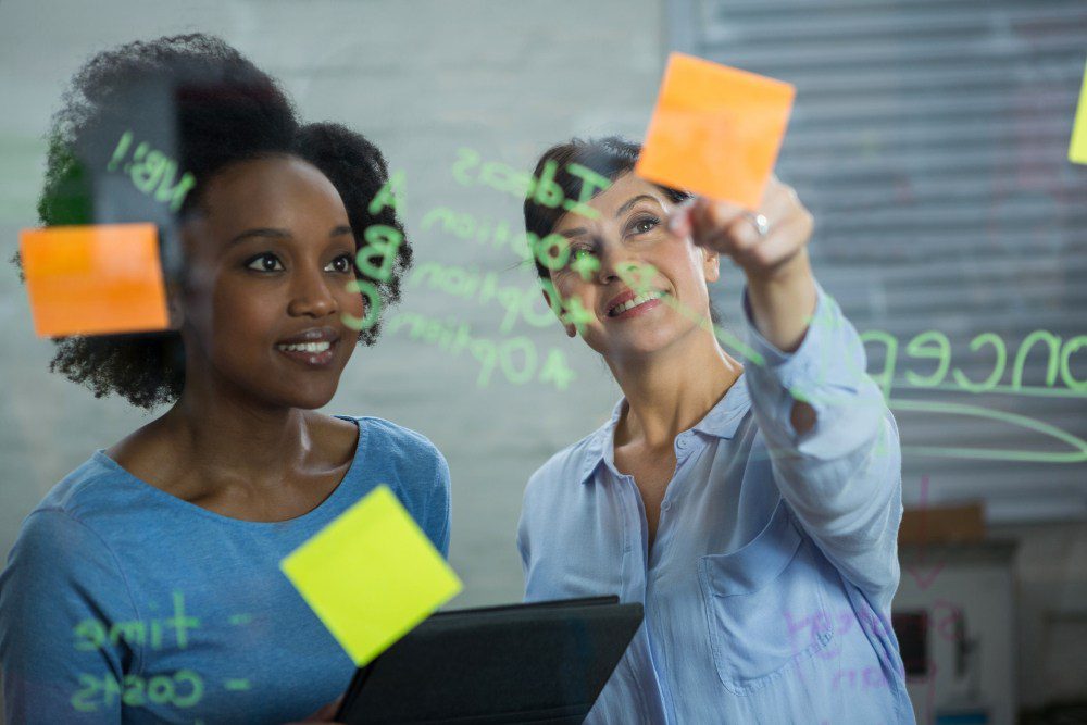 mulheres colam post-its em mural transparente fazendo capacitação sobre soft skills