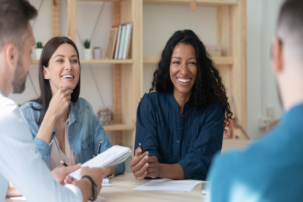 Feliz empresária afro-americana contando aos funcionários sobre o projeto sentado à mesa na sala de reuniões de coworking na reunião.