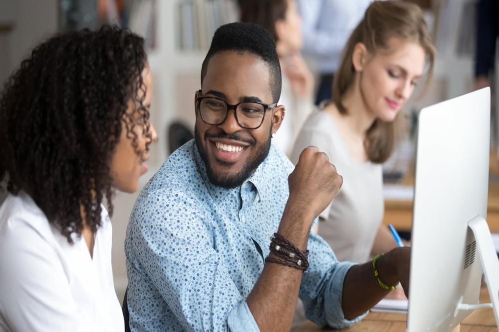 Funcionários que trabalham em escritório compartilhado, close em um homem negro sorridente conversando e sorrindo com um colega sentado à mesa usando o pc