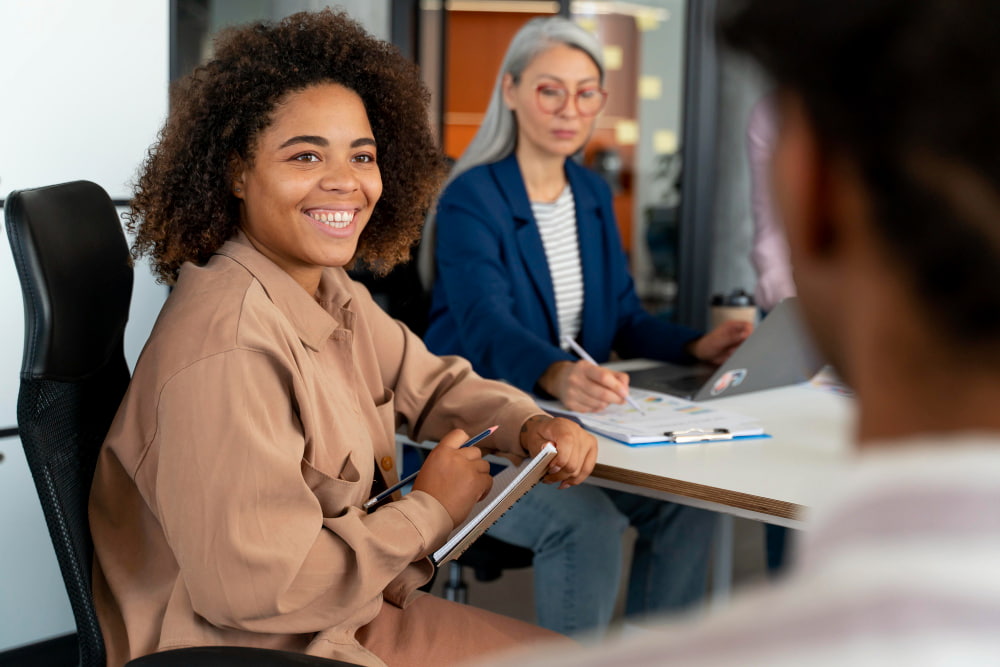 mulher negra sorridente sentada em uma mesa de reuniões com seus colegas de trabalho.